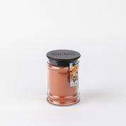 The Harvest Pumpkin 8oz Jar Candle by Bridgewater Candle Company features orange wax, a black lid, and a label with pumpkins and baked goods. This cozy autumn scent adds fall ambiance to any room. Shown against a plain white background.
