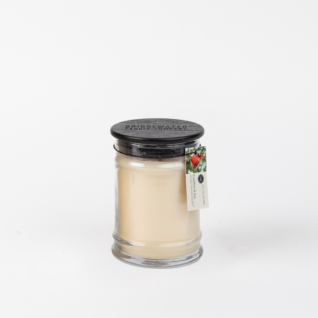 The Bridgewater Candle Company Comfort & Joy 8oz Small Jar Candle has ivory wax, a black embossed lid, and comes with a tag featuring strawberries and branding. The jar is set against a plain white background, evoking comfort and joy.