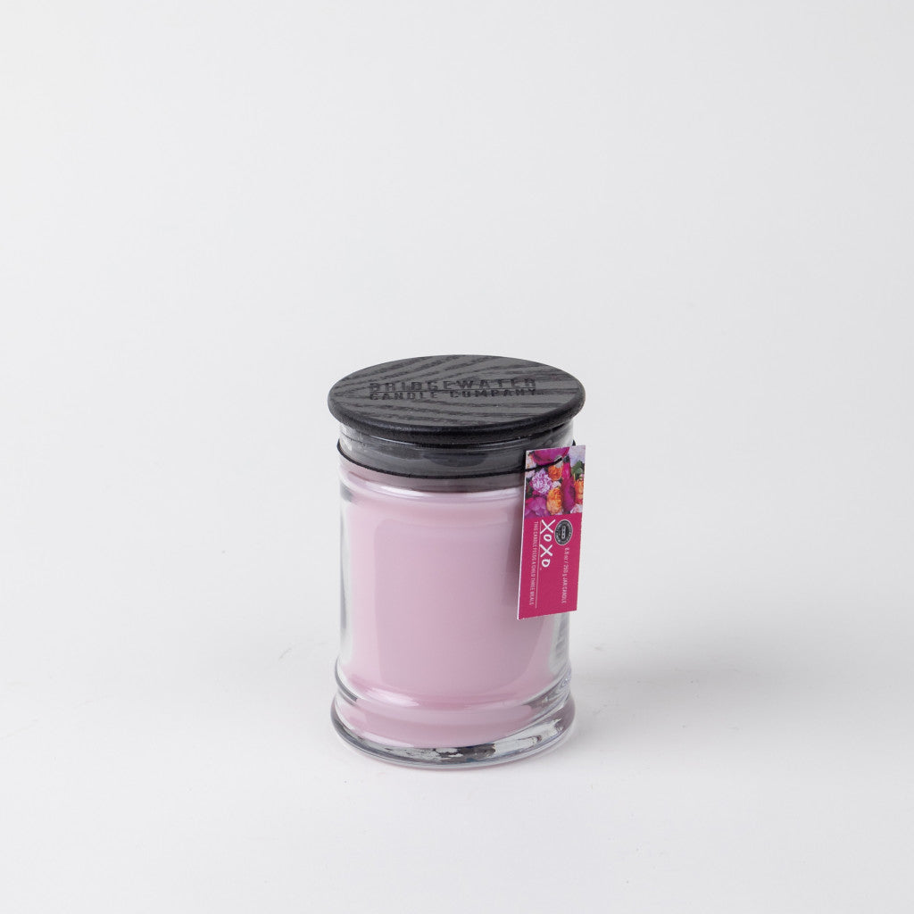 The Bridgewater Candle Company XOXO 8oz Small Jar Candle features light pink soy-blend wax, a black wooden lid, and a pink tag on top, all set against a plain white background.