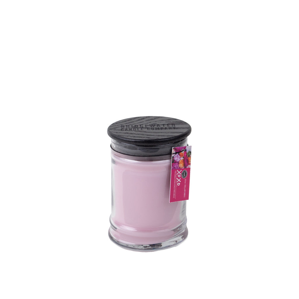 The Bridgewater Candle Company XOXO 8oz Small Jar Candle features light pink soy-blend wax, a dark lid, a colorful tag on top, and is infused with Bridgewater’s signature fragrance.