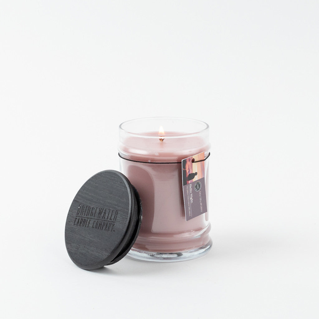 A lit pink Hello Night 8oz Jar Candle by Bridgewater Candle Company sits in a clear glass jar with a black lid, made from soy-blend wax and featuring a Bridgewater fragrance. A small tag is attached, all on a plain white background.