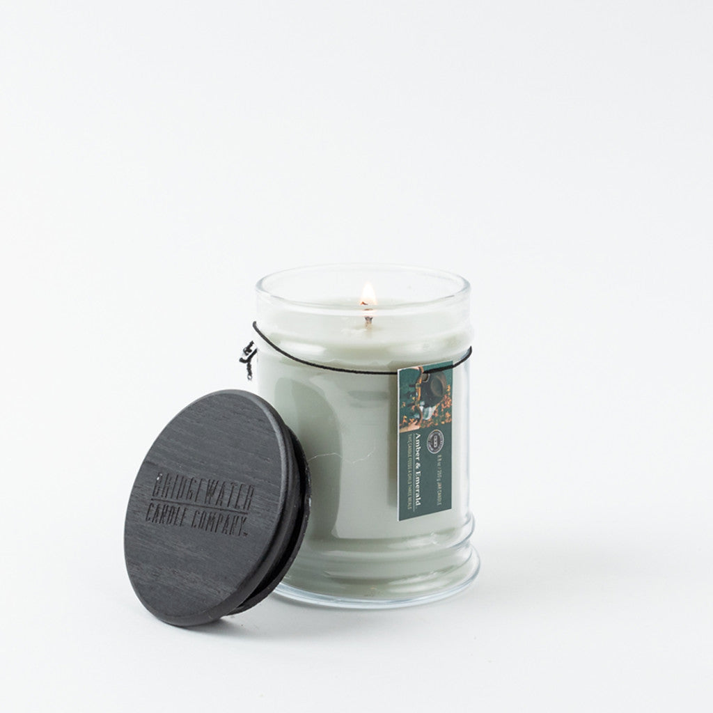 The Bridgewater Candle Company Amber & Emerald 8oz Jar Candle, made with soy-blend wax, burns in a clear glass jar with a black embossed lid nearby. A green label with text and an image decorates the jar against a plain white background.