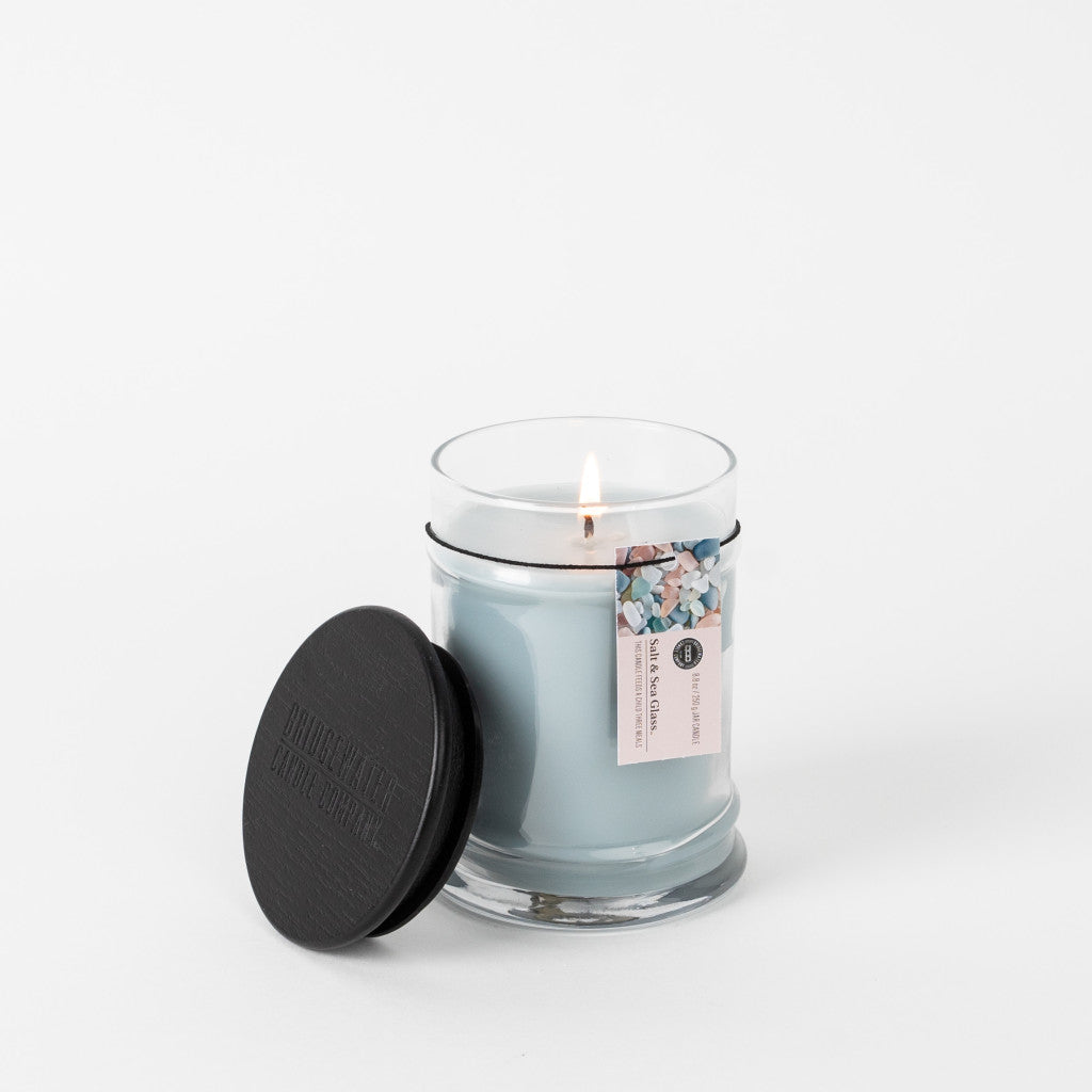 A glowing Salt & Sea Glass 8oz Small Jar Candle by Bridgewater Candle Company sits in a clear glass jar with a black lid nearby. The soy-blend wax has pastel bits on top, releasing a fresh scent against a white background.