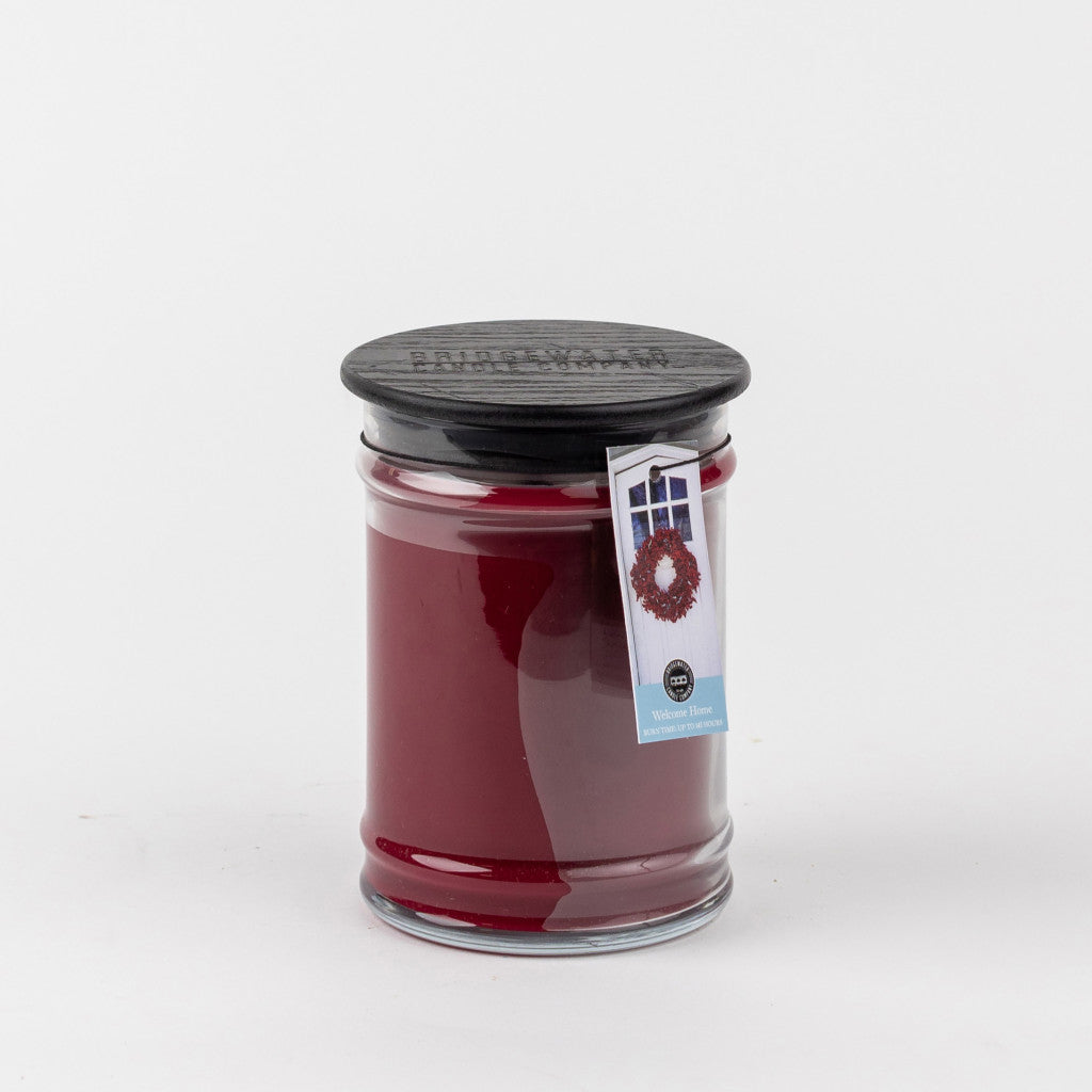 The Bridgewater Candle Company Welcome Home 18oz Large Jar Candle features a black lid, comes in a clear glass jar with a wreath tag, and hints at its inviting fragrance, all set against a plain white background.