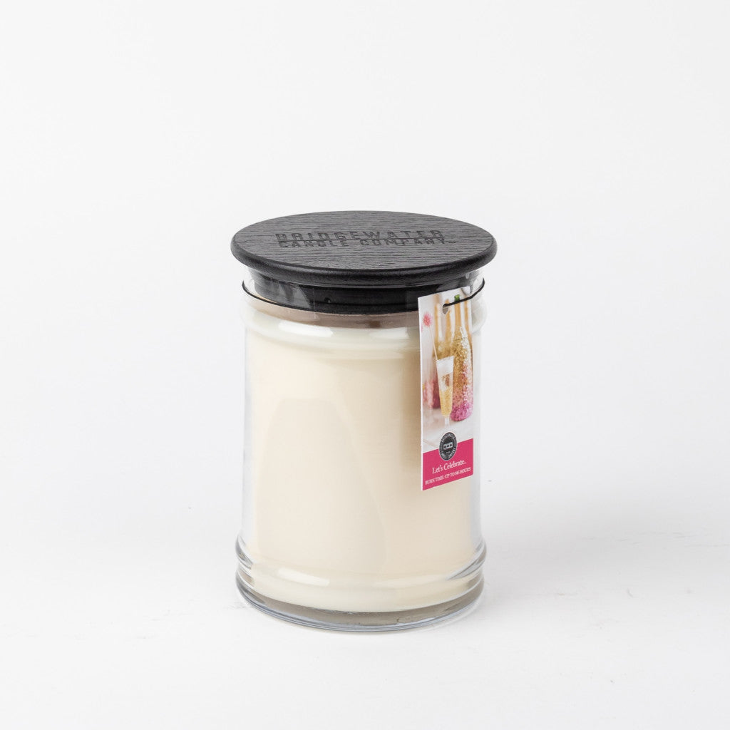 A white “Lets Celebrate” 18oz Large Jar Candle from Bridgewater Candle Company in a clear glass jar with a black wooden lid. A small label with pink accents and floral imagery hangs from the lid. The background is plain white.