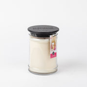 A white “Lets Celebrate” 18oz Large Jar Candle from Bridgewater Candle Company in a clear glass jar with a black wooden lid. A small label with pink accents and floral imagery hangs from the lid. The background is plain white.