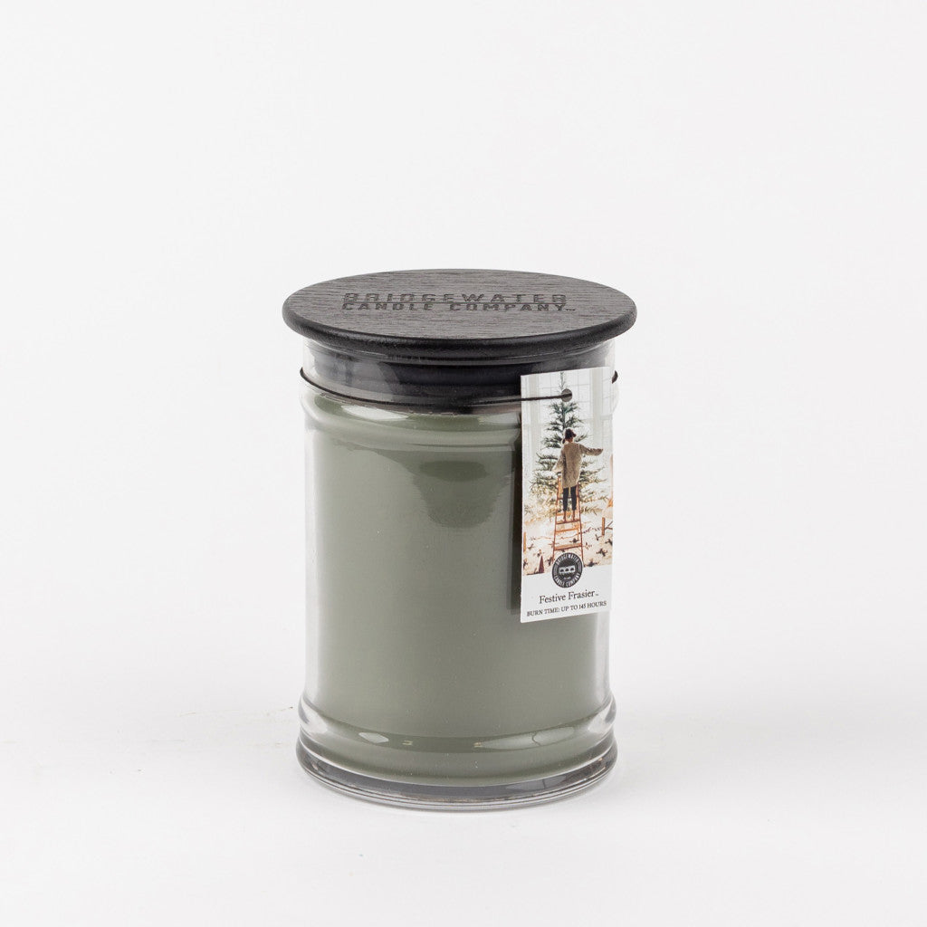 The Bridgewater Candle Company Festive Frasier 18oz Large Jar Candle features grayish-green wax, a black lid, and a pine tree label for a fresh, festive touch. Perfect for holiday ambiance. Shown on a plain white background.