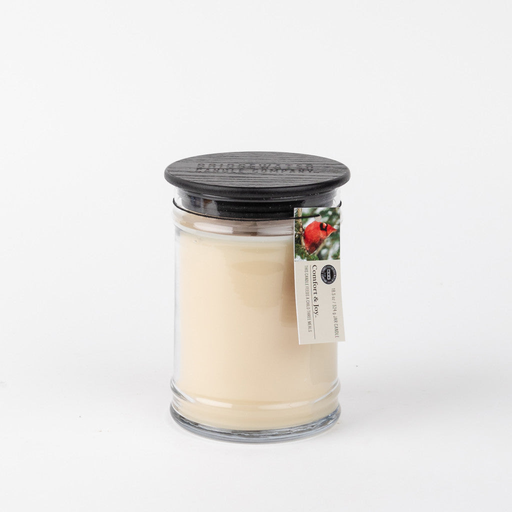 The Bridgewater Candle Company Comfort & Joy 18oz Large Jar Candle features creamy white wax, a black lid topped with a tag showing a red bird and green leaves, all set against a plain white background.