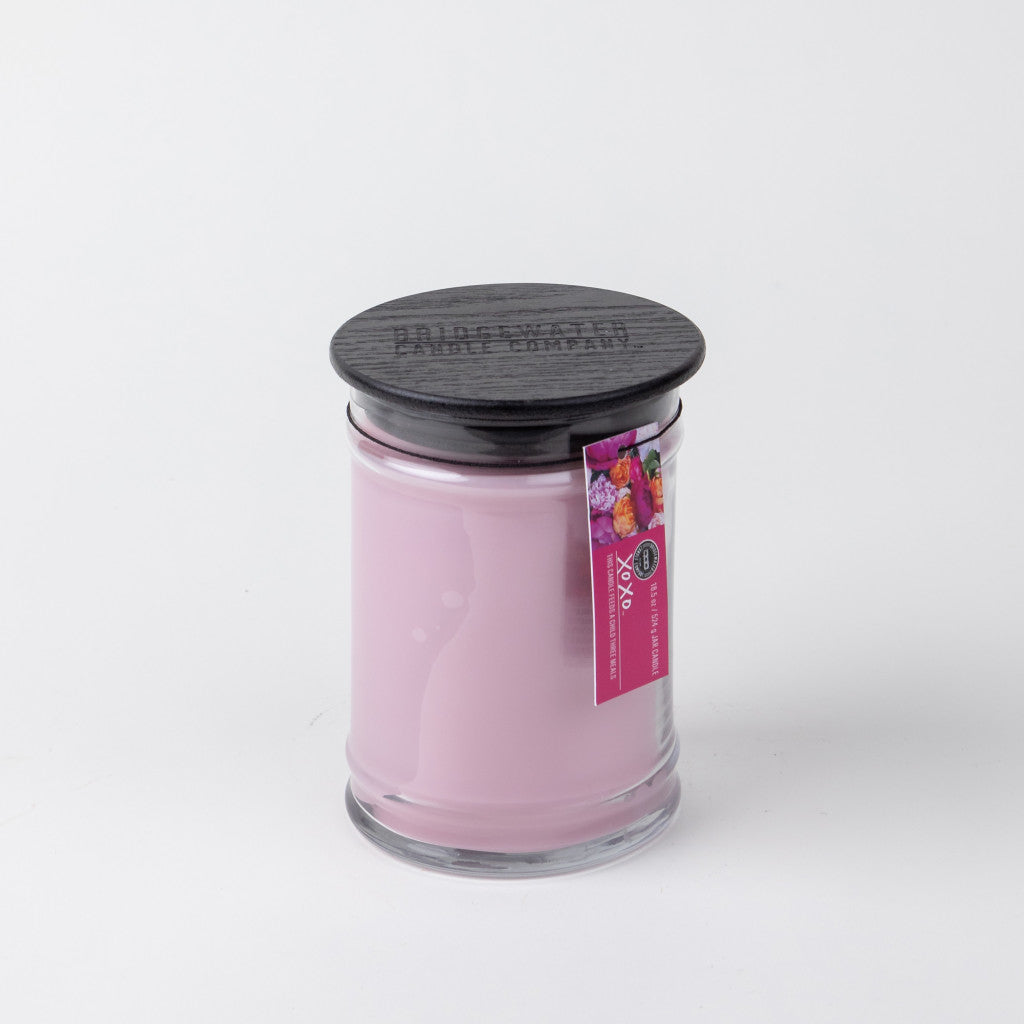 The XOXO 18oz Large Jar Candle by Bridgewater Candle Company features soy-blend wax in a light pink hue, a dark wooden lid, and a pink floral label with text, displayed against a plain white background.