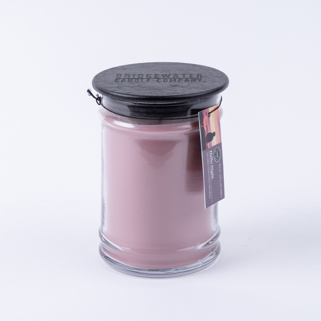 The Hello Night 18oz Jar Candle by Bridgewater Candle Company features pink soy-blend wax in a clear glass jar with black embossed lid and hanging tag showing branding and product details. Background is plain white.