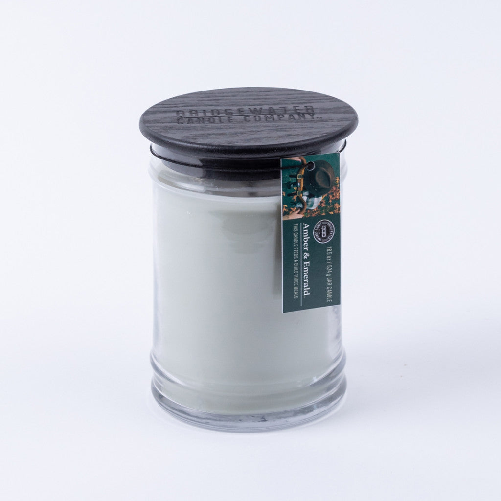 The Bridgewater Candle Company Amber & Emerald 18oz Large Jar Candle features soy-blend wax in a clear glass jar with a dark wooden lid and a green label, set against a plain white background.