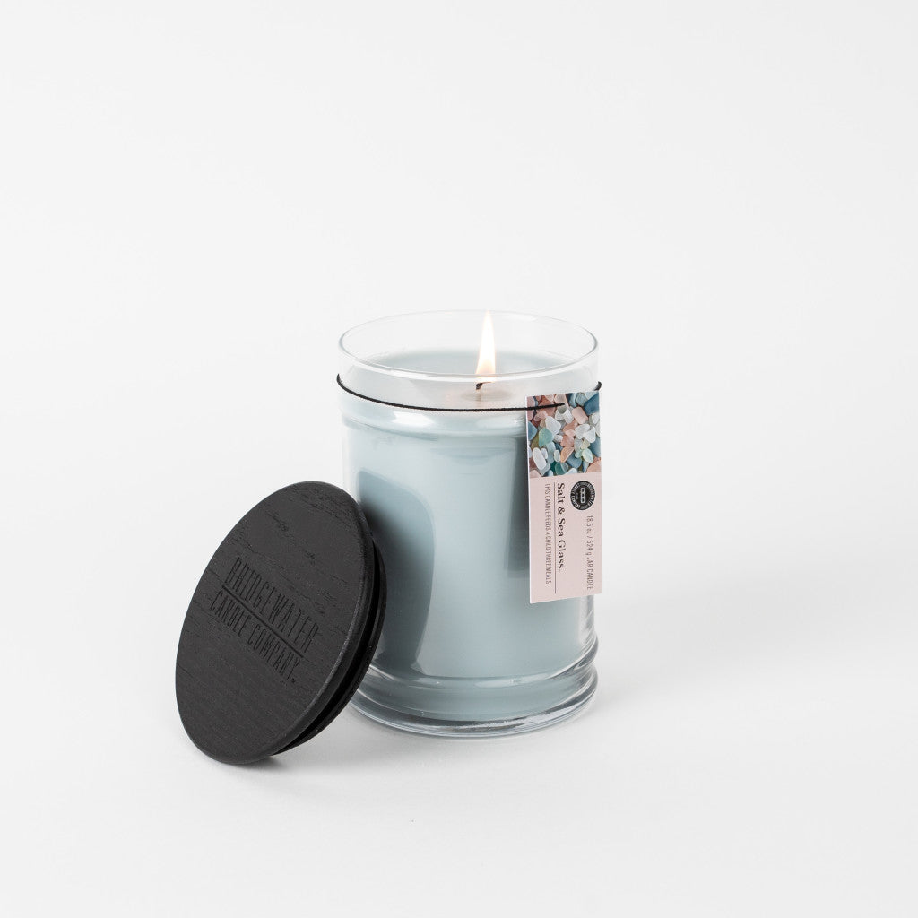 A Bridgewater Candle Company Salt & Sea Glass 18 oz Large Jar Candle, made with a soy-blend wax, burns in a clear jar with a black lid beside it. A small floral tag is attached to the jar. The background is white.
