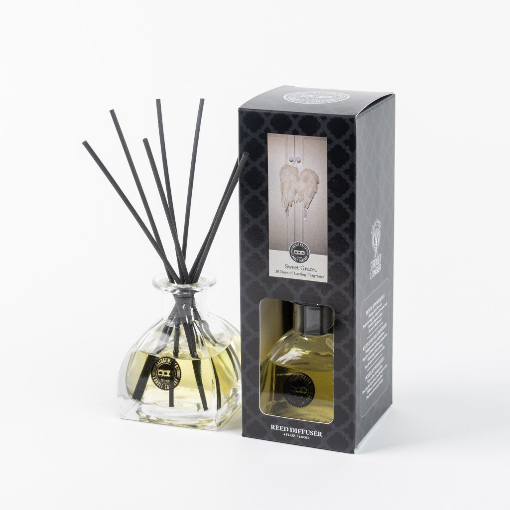 The Bridgewater Candle Company Sweet Grace Reed Diffuser features black reeds in a clear bottle with yellow liquid and comes in a black patterned box with angel wings, filling your space with an uplifting home fragrance.