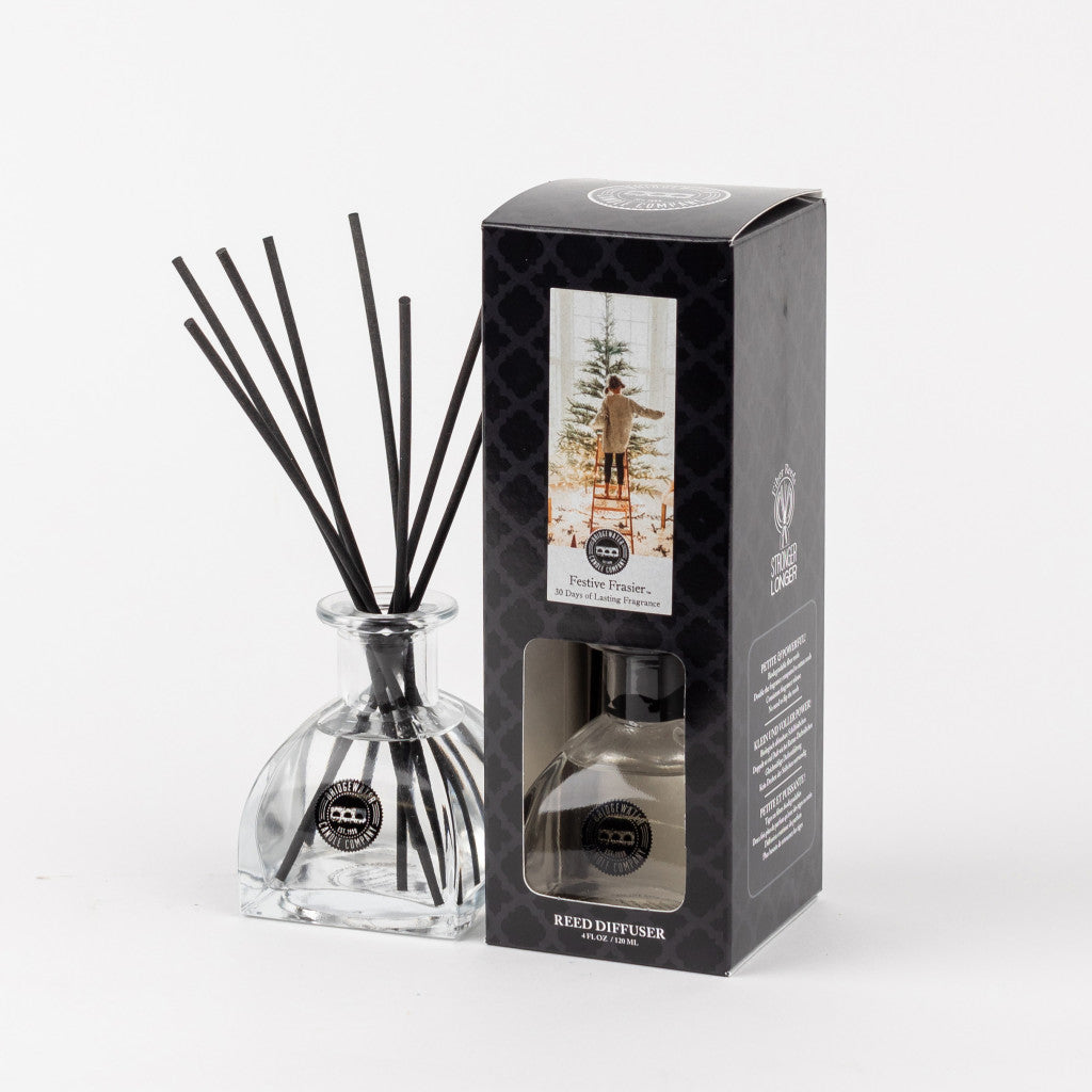 A Bridgewater Candle Company Festive Frasier Reed Diffuser with black fiber reeds stands by its box, which features a person in a snowy forest and describes the Festive Frasier fragrance.