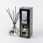 The Bridgewater Candle Company Lavender Fields Reed Diffuser, with fiber reeds and clear liquid, is displayed beside its black box packaging featuring lavender flower imagery and the “Lavender Fields” label.