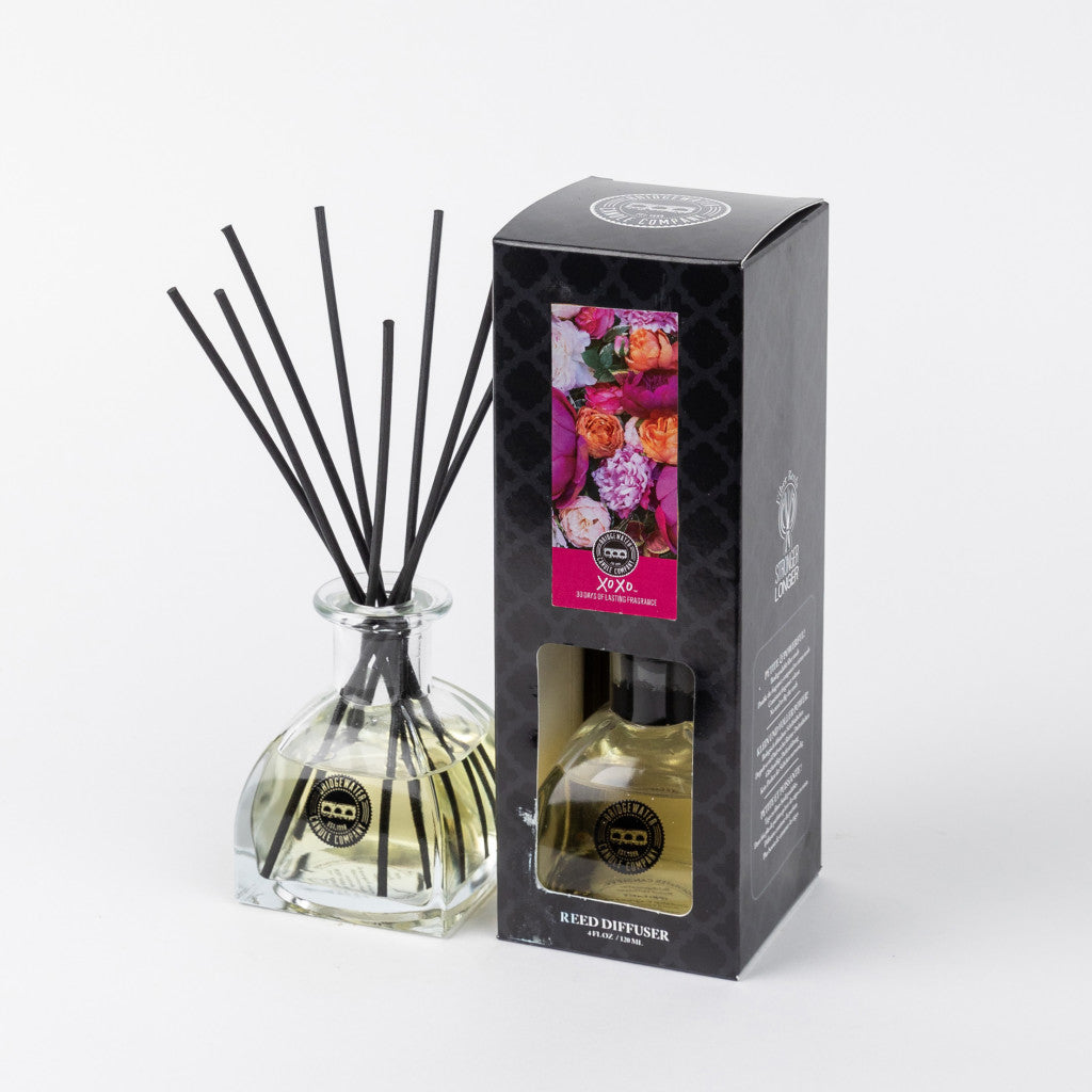 The XOXO Reed Diffuser by Bridgewater Candle Company features a glass bottle with black fiber reeds, paired with sleek black packaging adorned with colorful flowers and a window showcasing the premium fragrance inside.
