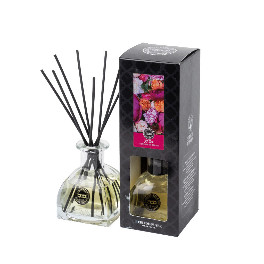 The Bridgewater Candle Company XOXO Reed Diffuser with black fiber reeds stands next to its stylish box, adorned with a floral design and the label Reed Diffuser. The premium diffuser contains a pale yellow fragrance liquid.