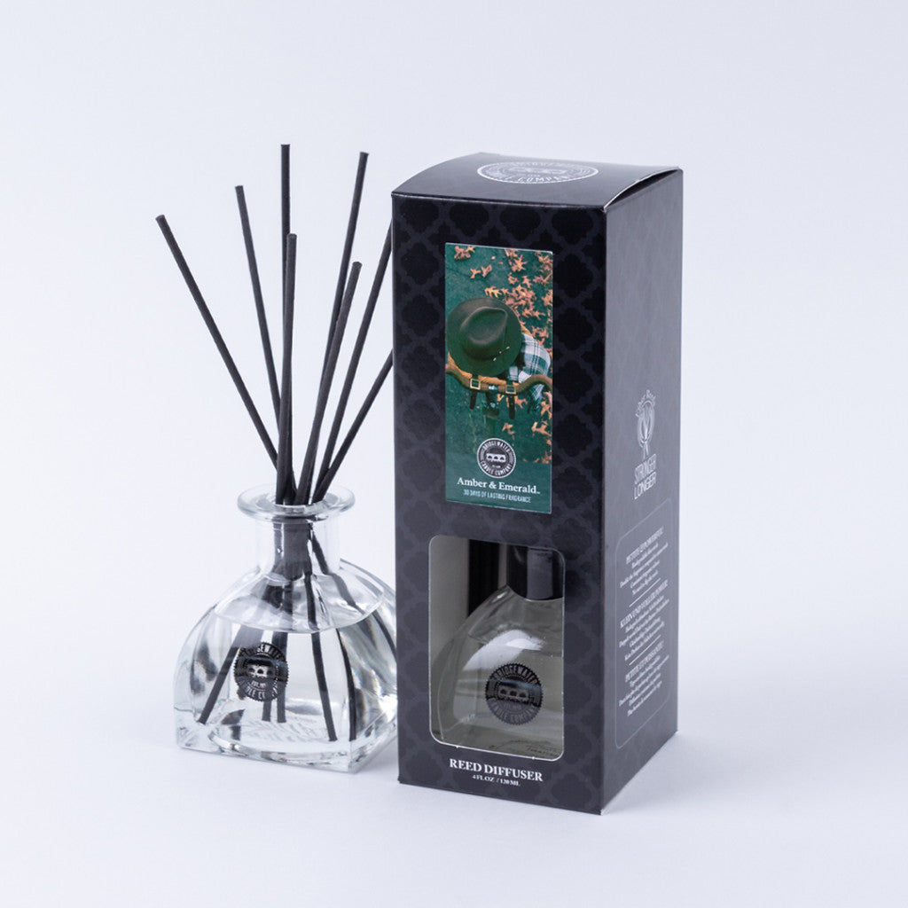 A Bridgewater Candle Company Amber & Emerald Reed Diffuser with black fiber reeds is displayed beside its black box, which features a green and gold design, a window revealing the premium fragrance bottle, and elegant branding.