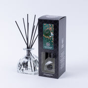 A Bridgewater Candle Company Amber & Emerald Reed Diffuser with black fiber reeds is displayed beside its black box, which features a green and gold design, a window revealing the premium fragrance bottle, and elegant branding.
