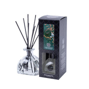 A glass Bridgewater Candle Company Amber & Emerald Reed Diffuser with black reeds stands beside a black box featuring a green design, a transparent window displaying the premium fragrance inside, and the product name.