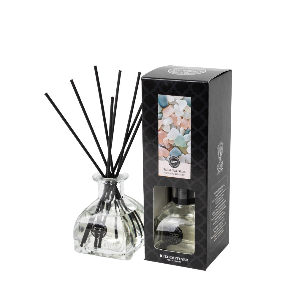 The Bridgewater Candle Company Salt & Sea Glass Reed Diffuser stands next to its black box, which features pastel stone designs and a cutout window that reveals the stylish home fragrance bottle inside.