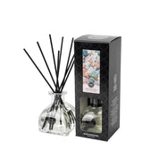 The Bridgewater Candle Company Salt & Sea Glass Reed Diffuser stands next to its black box, which features pastel stone designs and a cutout window that reveals the stylish home fragrance bottle inside.