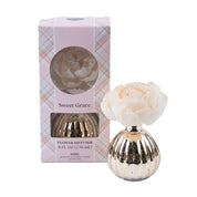 The Sweet Grace Mercury Flower Diffuser by Bridgewater Candle Company features a white, flower-shaped diffuser in a gold ribbed vase beside its pink plaid box, adding elegant fragrance and style to your space.