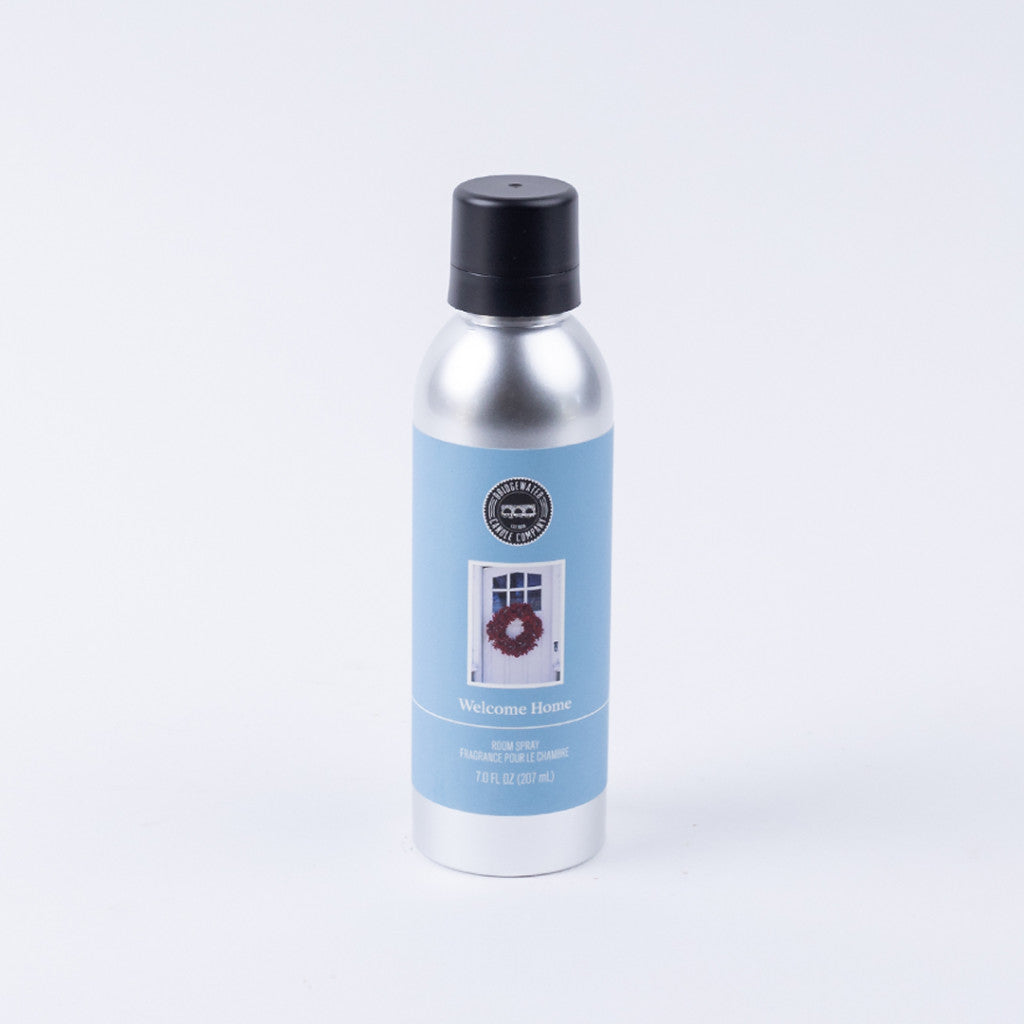 The Bridgewater Candle Company Welcome Home Non-Aerosol Room Spray features a silver can with a black cap and blue label with a door and wreath image. The eco-friendly spray stands upright against a plain white background.