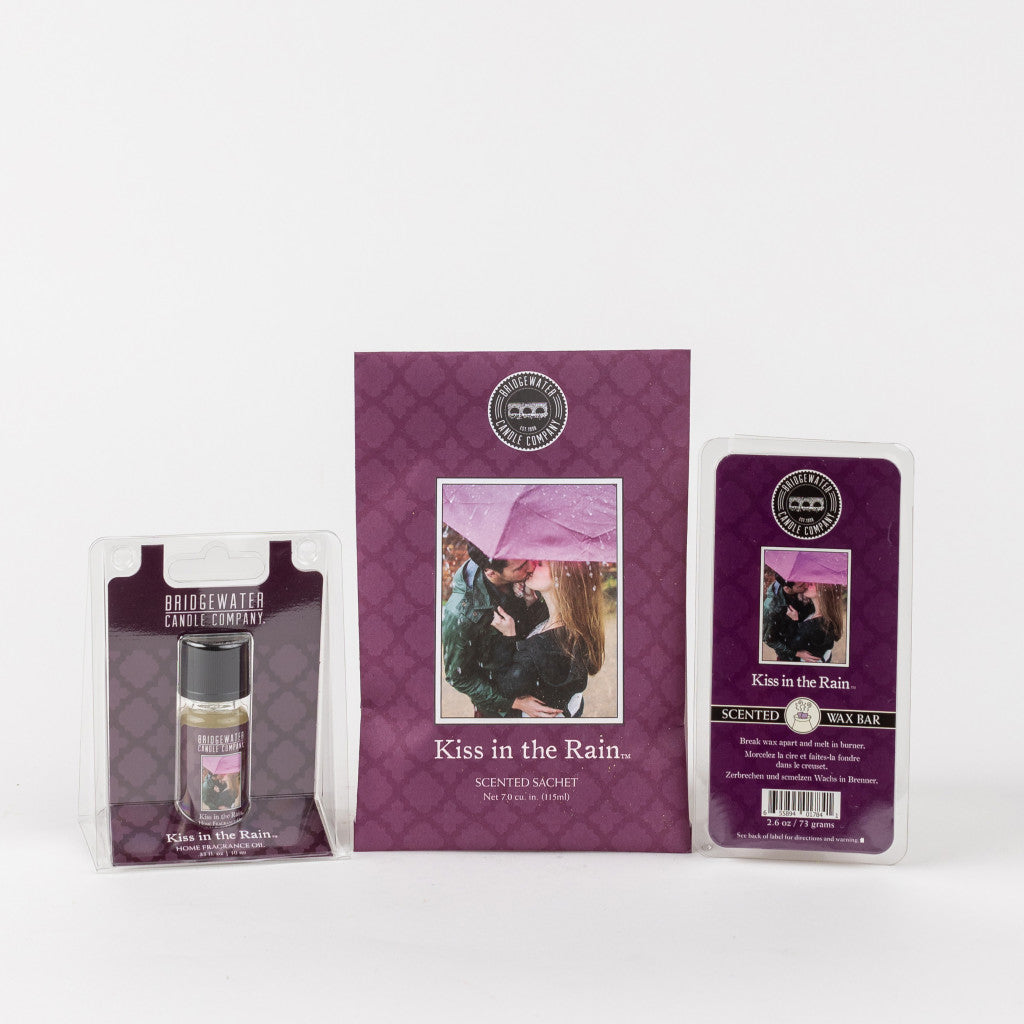 The Bridgewater Candle Company Kiss in the Rain Flameless Bundle includes a home fragrance oil, scented sachet, and wax bar in purple packaging with a couple under a pink umbrella, bringing romantic ambiance to your space.