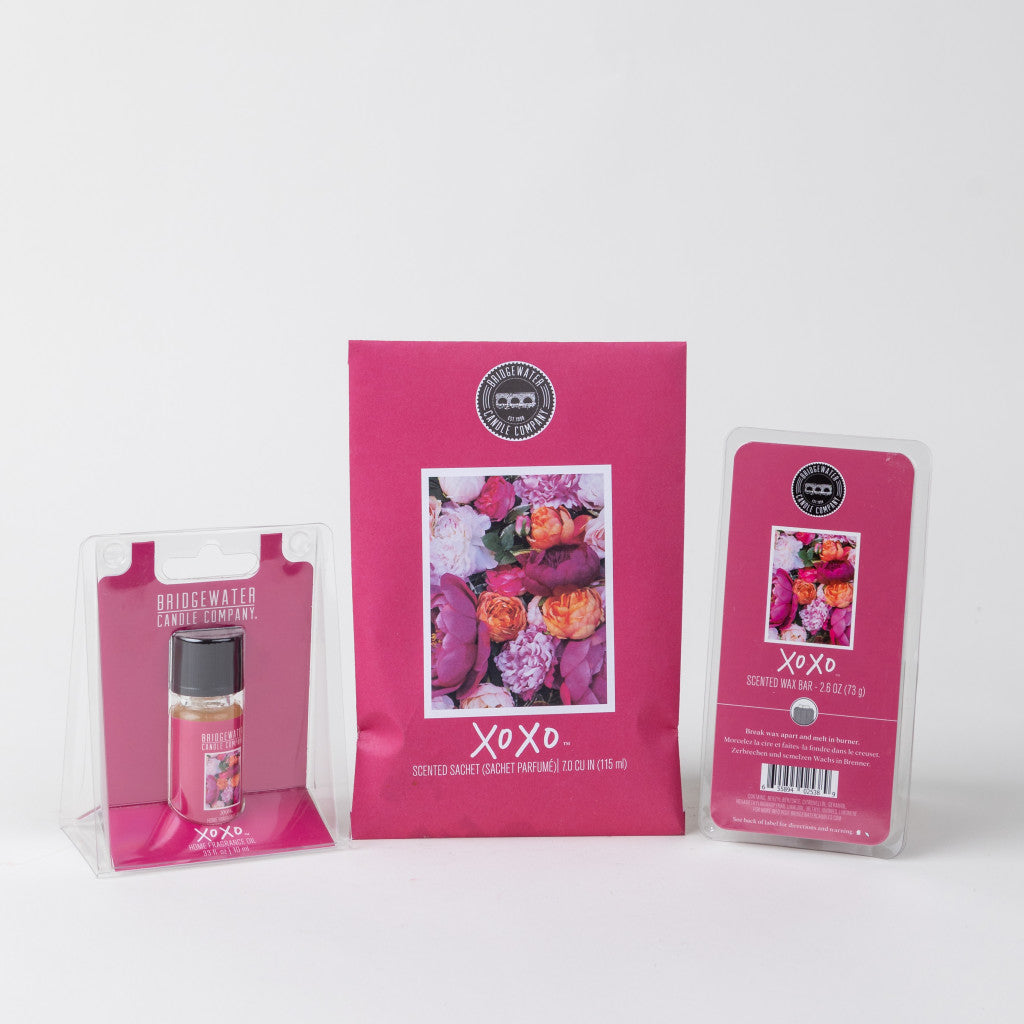 The Bridgewater Candle Company XOXO Flameless Bundle includes a home fragrance oil, scented sachet, and soy-blend wax bar, all in pink packaging with floral designs featuring pink, white, and orange blossoms.