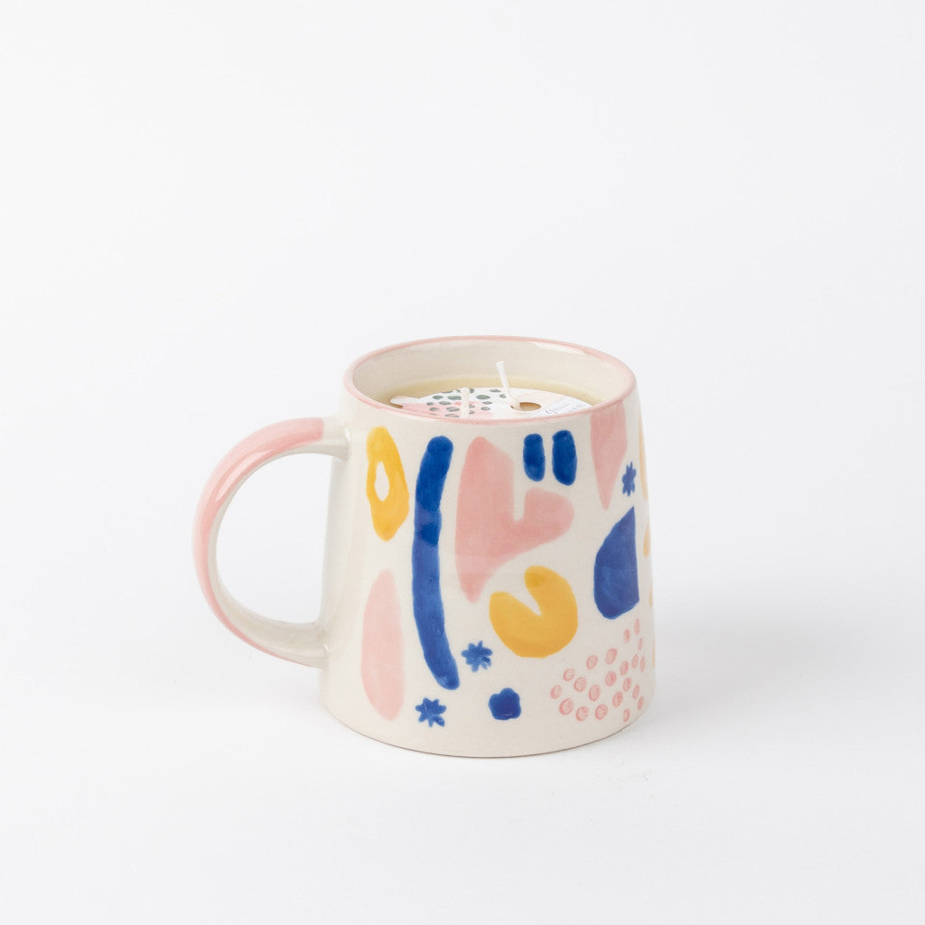 A Sweet Grace Collection Candle #047 by Bridgewater Candle Company, made from soy-blend wax, sits in a ceramic mug with a pink handle and abstract blue, yellow, and pink shapes on a plain white background.