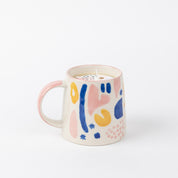 A Sweet Grace Collection Candle #047 by Bridgewater Candle Company, made from soy-blend wax, sits in a ceramic mug with a pink handle and abstract blue, yellow, and pink shapes on a plain white background.