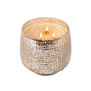 A lit Bridgewater Candle Company Sweet Grace Collection Candle #051, made with soy-blend wax, glows in a textured metallic gold and silver jar against a white background. Experience this best-selling fragrance’s elegant allure.