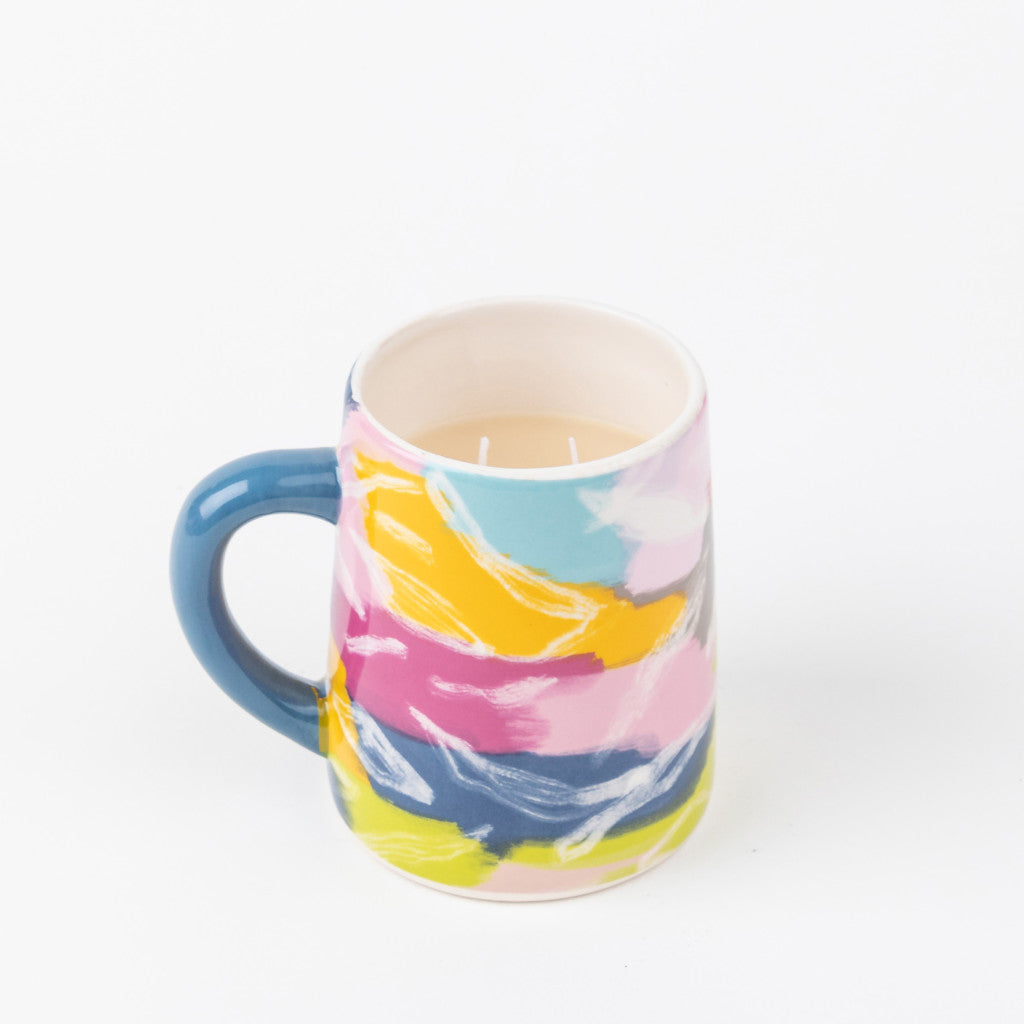 A ceramic mug with a blue handle and a vibrant abstract design in pink, yellow, blue, and white holds a light liquid, evoking the calming scent of Bridgewater Candle Companys Sweet Grace Collection Candle #055. Set against a plain white background.
