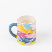 A ceramic mug with a blue handle and a vibrant abstract design in pink, yellow, blue, and white holds a light liquid, evoking the calming scent of Bridgewater Candle Companys Sweet Grace Collection Candle #055. Set against a plain white background.