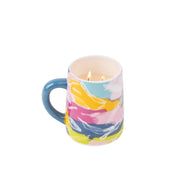 A colorful ceramic mug with a blue handle and abstract patterns holds the Sweet Grace Collection Candle #055 by Bridgewater Candle Company, featuring soy-blend wax and two visible flames.