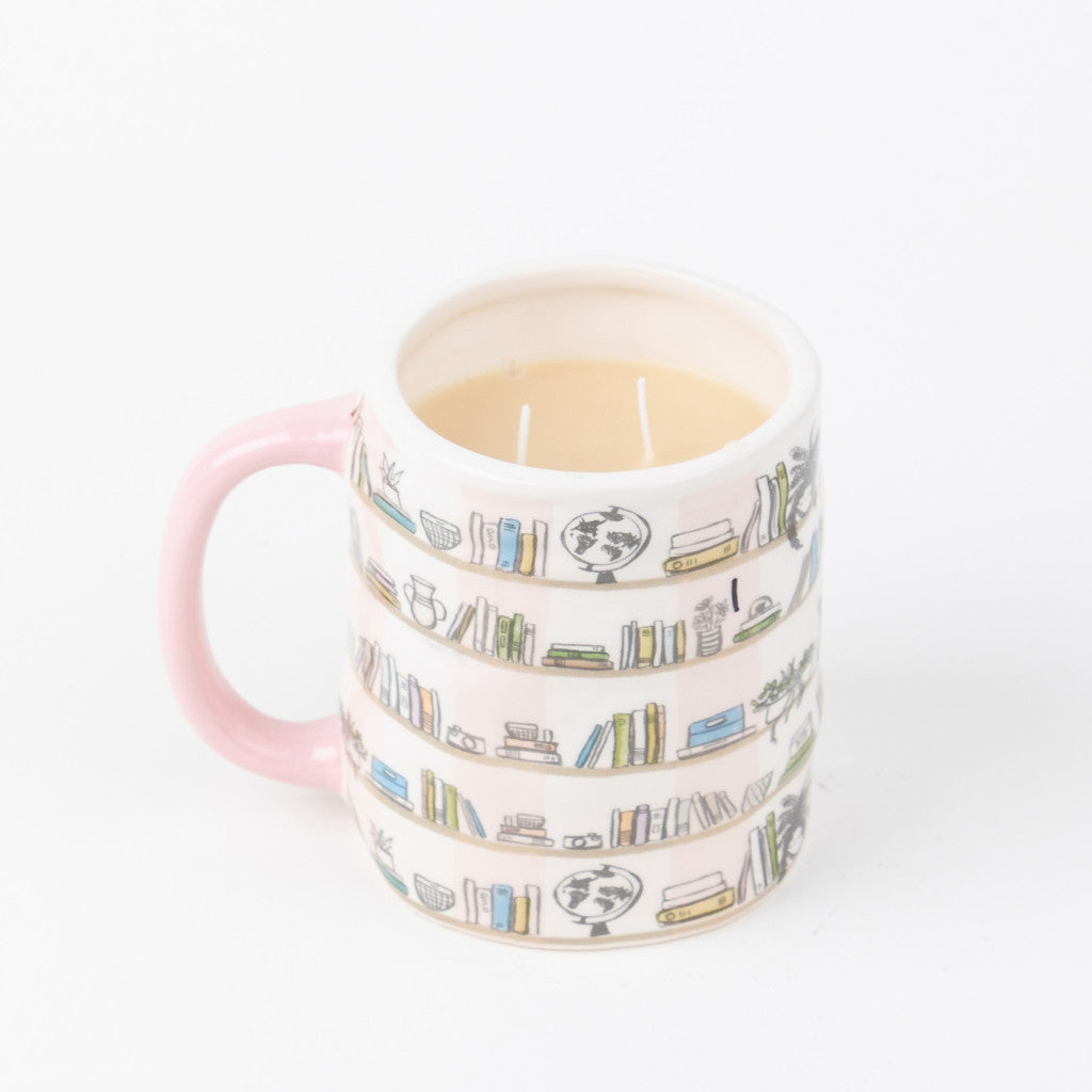 A white mug with a pink handle, decorated with bookshelf illustrations, holds the Sweet Grace Collection Candle #056 by Bridgewater Candle Company—a creamy, two-wick candle made from soy-blend wax. Plain white background.