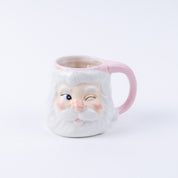 The Bridgewater Candle Company Sweet Grace Santa Mug is a ceramic reusable mug shaped like a winking Santa with a white beard, rosy cheeks, and a pink handle—ideal for holiday candle lovers or as festive decor.