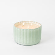 A light green, ribbed ceramic container holds the Bridgewater Candle Company Sweet Grace Collection Candle #065 with soy-blend wax, its three flames glowing brightly against a plain white background.