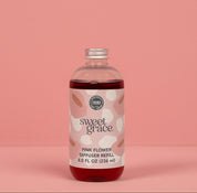 A clear bottle of Bridgewater Candle's Sweet Grace Pink Dye Fragrance Refill Oil, filled with dark pink liquid, stands against a solid pink background. Its label displays abstract pink and beige patterns, reflecting our best-selling scent.