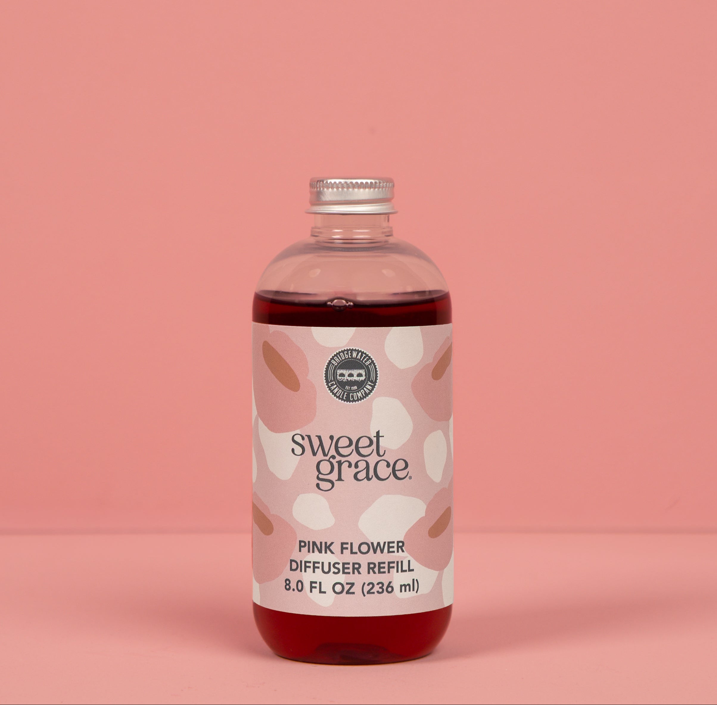 A clear bottle of Bridgewater Candle's Sweet Grace Pink Dye Fragrance Refill Oil, filled with dark pink liquid, stands against a solid pink background. Its label displays abstract pink and beige patterns, reflecting our best-selling scent.