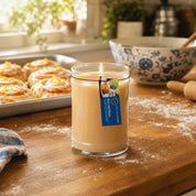 A lit Rise & Shine 18oz Large Jar Candle by Bridgewater Candle, made with soy-blend wax, sits on a floured wooden kitchen counter near pastries, a rolling pin, and a blue patterned bowl as sunlight streams through the window.
