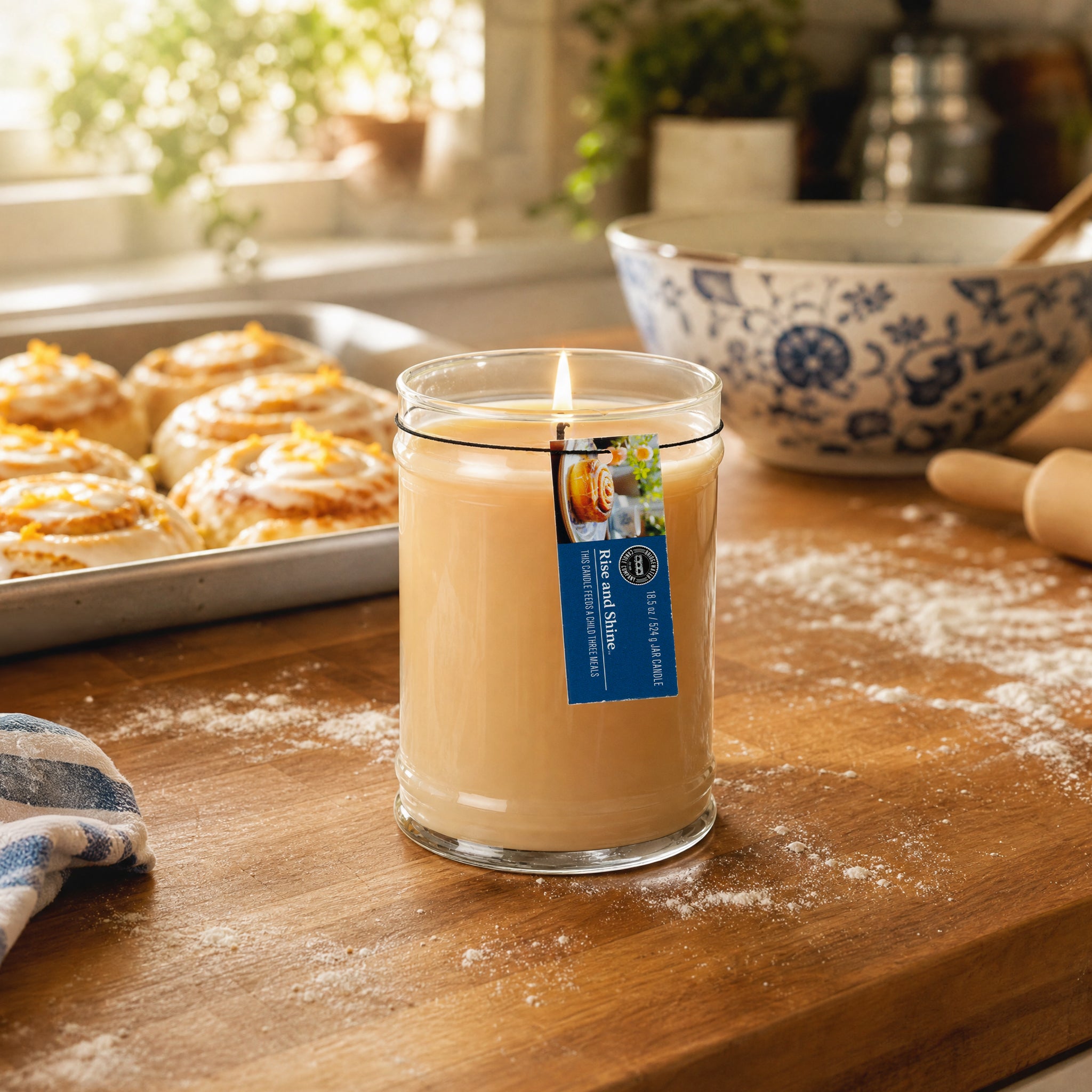 A lit Rise & Shine 18oz Large Jar Candle by Bridgewater Candle, made with soy-blend wax, sits on a floured wooden kitchen counter near pastries, a rolling pin, and a blue patterned bowl as sunlight streams through the window.