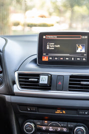 A small square Auto Vent Clip-Laundry Line by Bridgewater Candle clipped to the center car vent, with a vehicle dashboard display visible above showing music playback.