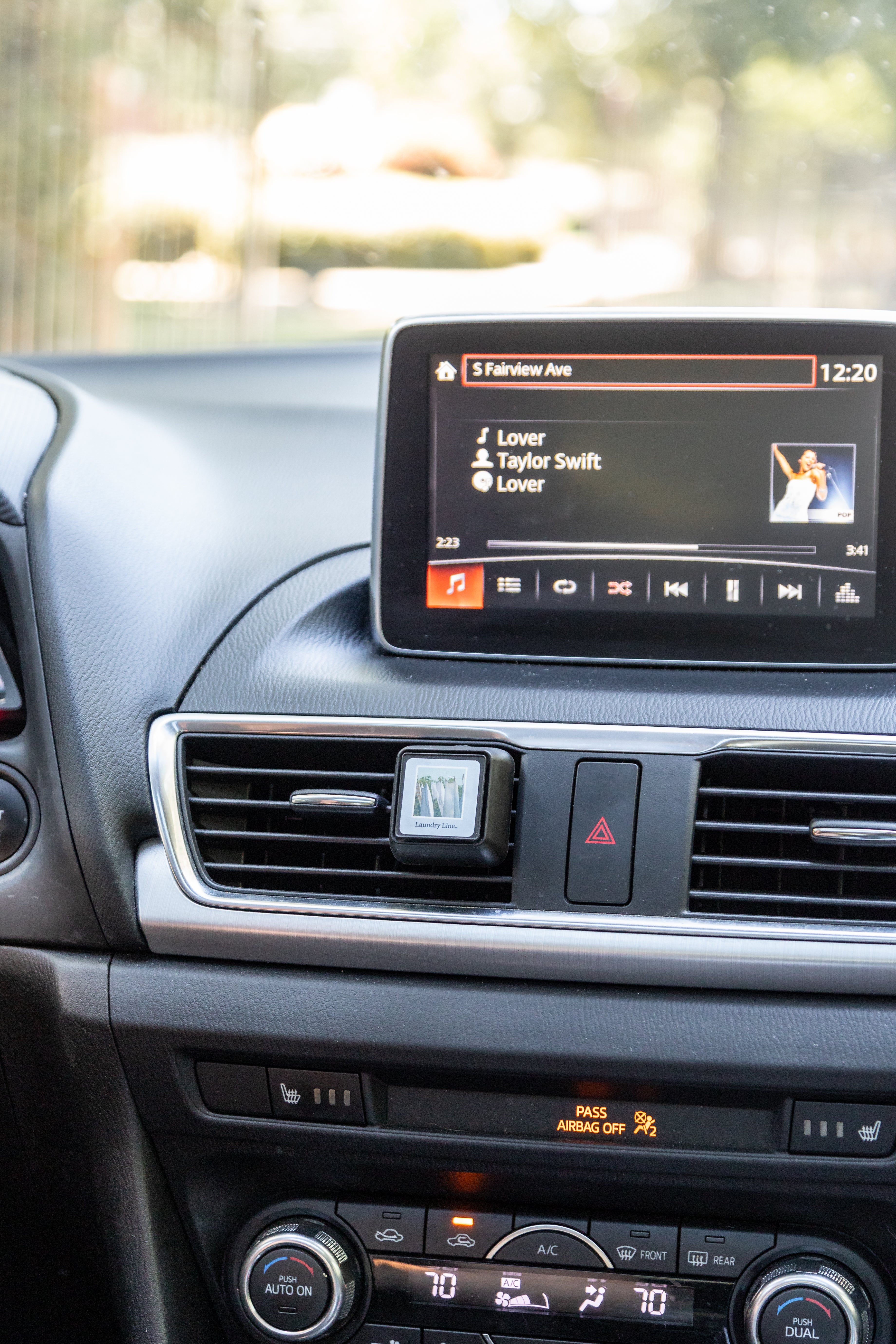 A small square Auto Vent Clip-Laundry Line by Bridgewater Candle clipped to the center car vent, with a vehicle dashboard display visible above showing music playback.