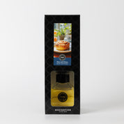The Bridgewater Candle Rise & Shine Reed Diffuser features yellow fragrance liquid in a clear bottle, visible through a window in its black patterned box displaying a cinnamon roll and coffee image on the packaging.