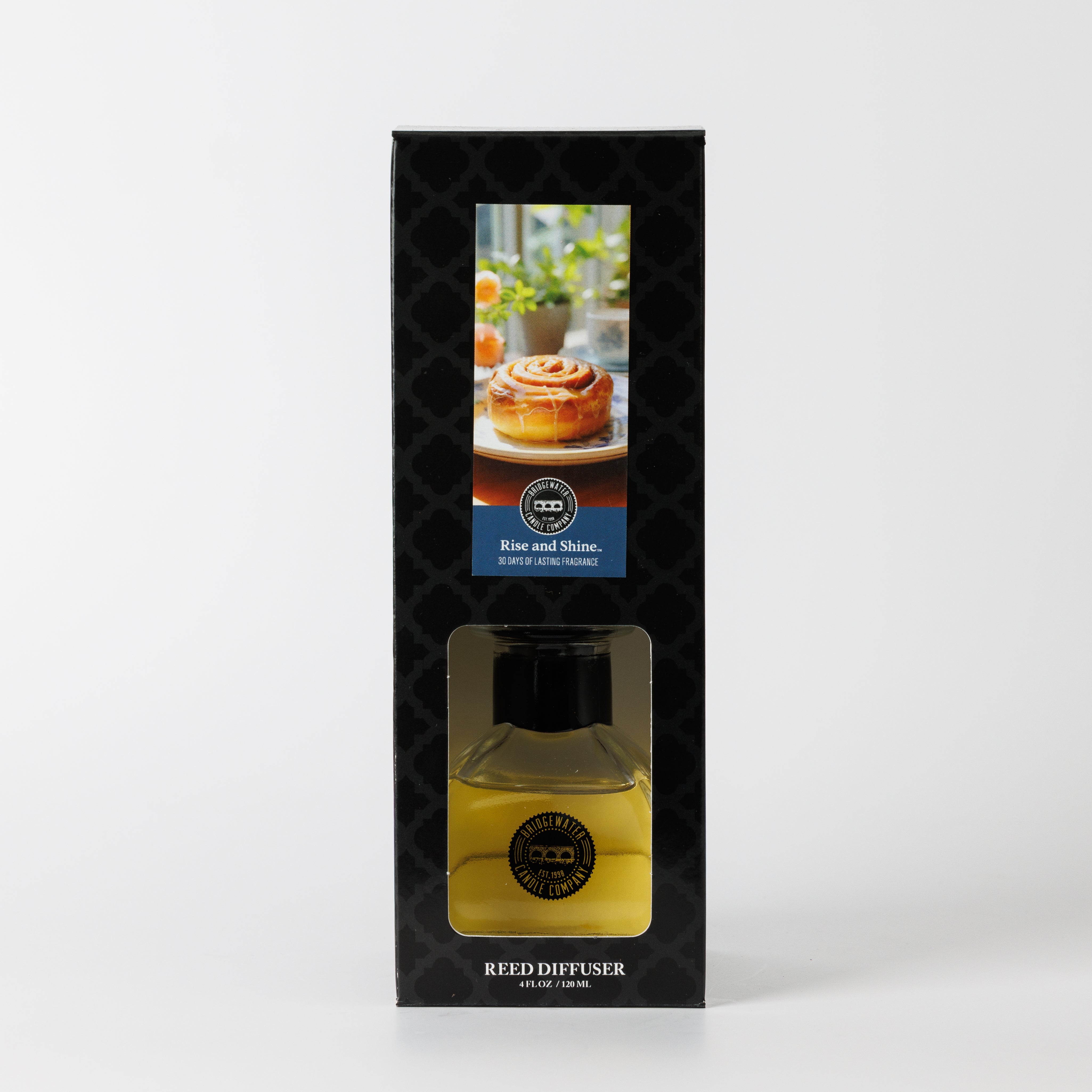 The Bridgewater Candle Rise & Shine Reed Diffuser features yellow fragrance liquid in a clear bottle, visible through a window in its black patterned box displaying a cinnamon roll and coffee image on the packaging.