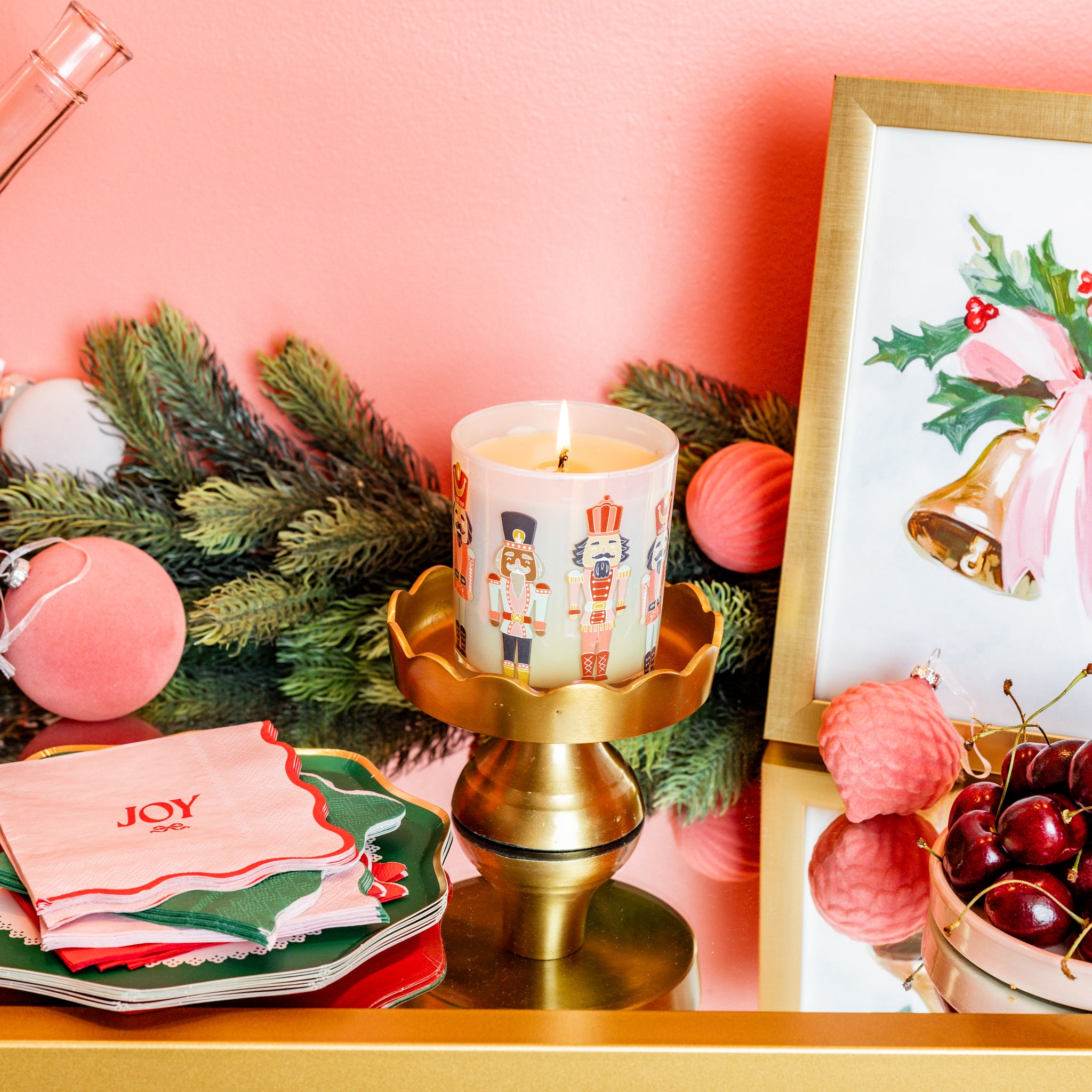 A Bridgewater Candle Sweet Grace Holiday Nutcracker Candle in a festive holder sits on a gold tray with pine branches, pink ornaments, a JOY holiday napkin set, and a framed holly and bells print against a pink wall.