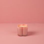 A pastel pink Sweet Grace Collection Candle #075 by Bridgewater Candle, in a flower-shaped ribbed glass vessel, rests on a matching pink surface and background.