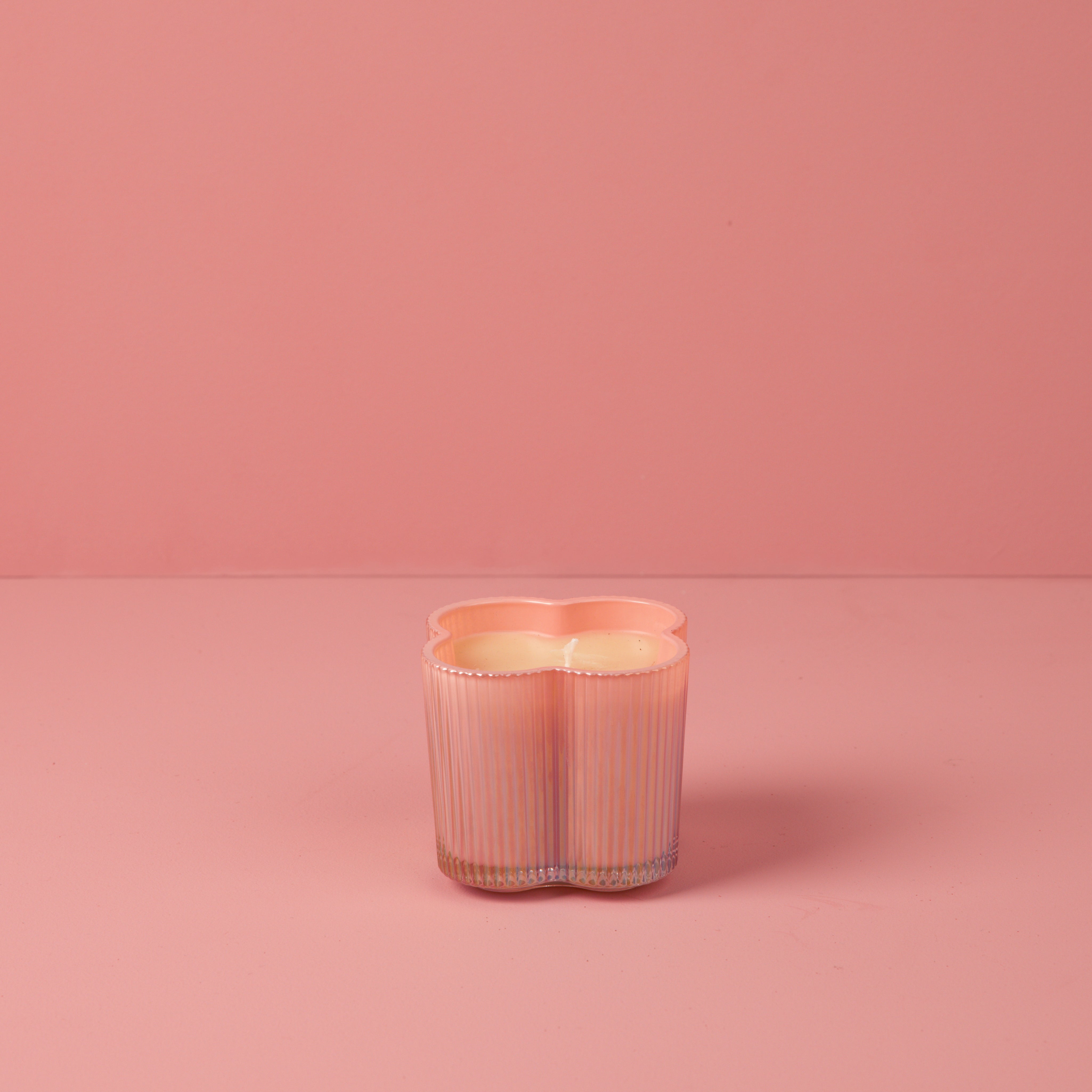A pastel pink Sweet Grace Collection Candle #075 by Bridgewater Candle, in a flower-shaped ribbed glass vessel, rests on a matching pink surface and background.